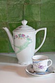 Coffee pot, cup and saucer with floral pattern in front of green wall tiles