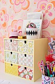 Miniature chest of drawers with floral wallpaper on front and pot of writing utensils against retro floral wallpaper