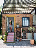 Frau in Tür mit Blumentopf in Hand, davor Aussentreppe mit gestreiftem Teppichläufer, Landhaus mit Ziegelfassade