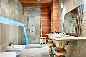 Bad mit bläulichem Leuchtband zwischen Natursteinfliesen und horizontaler Holzverkleidung, Waschtisch mit geschnitzten Holzstützen und gerahmtem Spiegel