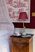 Table lamp with red fabric lampshade on bedside cabinet against red and white toile de jouy wallpaper