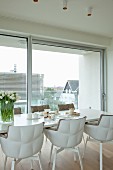 Weißer Esstisch mit zwei Frühstücksgedecken, weißem Tulpenstrauß und gepolsterten Designerstühlen vor Fensterfront