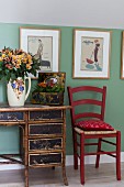 Roter Küchenstuhl neben antikem Schreibtisch mit Bambusgestell vor pastellgrüner Wand, Bilder mit Retro Motiven