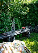 Shady seating area in garden with weathered wooden bench and patterned picnic blanket in foreground