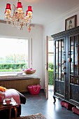 Dark, painted glass-fronted wooden cabinet in rustic interior with small red lampshades on chandelier