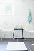 White cord chairs flanking black delicate side table and white bathmat on grey tiled floor