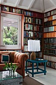 Seating area with caramel-coloured sofa, wicker trunk and petrol blue table in front of full-wall bookcases