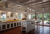Long kitchen counter and masonry breakfast bar with wire stools below white-painted, wood-beamed ceiling