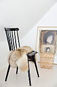 Animal-skin rug on black-painted wooden chair in corner below sloping ceiling; stool in front of painting leaning against wall in background