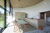 Classic chairs at delicate, long wooden table between masonry kitchen counter with cupboards and glass façade in open-plan interior