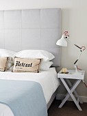 Bed with pale grey headboard next to desk lamp on folding side table