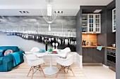 Tulip table, Eames Plastic Armchairs and blue sofa in front of mural wallpaper; open-plan, grey designer kitchen to one side