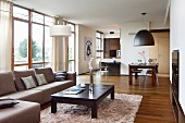 Offener Wohnbereich mit Sofa und Coffeetable aus dunklem Holz, im Hintergrund Essplatz vor Wandscheibe zur Designerküche