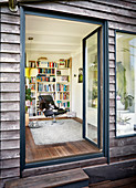 Holzhaus mit offener Terrassentür und Blick in Wohnraum auf Ledersessel vor Bücherwand