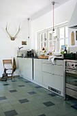 Modern kitchen counter below window and antlers hung on wall of rustic kitchen; tiled floor with dark accent tiles