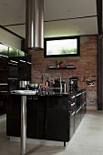 Black free-standing designer kitchen counter below cylindrical stainless steel extractor hood in front of brick wall