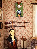 Painted figurine in corner below hunting rifles on wall with floral wallpaper