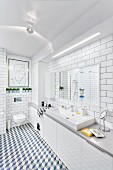 Modern, white bathroom with tiled walls & floor, long washstand with base cabinets & strip light over mirror; modern artwork above wall-mounted toilet in background
