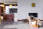 Retro-style, wood-framed armchair in front of fireplace, grey stone floor, kitchen counter and white fitted cabinets in open-plan kitchen to one side
