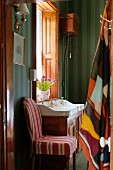 Chair with striped cover against dark green striped wallpaper in bathroom with antique-style washstand below window