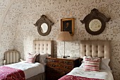 Twin beds with button-tufted headboards, antique chest of drawers, round mirrors with wooden frames and floral wallpaper in attic bedroom in English manor house