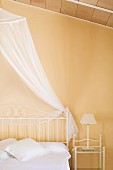 Bed with delicate, white metal frame and airy canopy in bedroom with pastel yellow wall