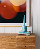 Light blue vase on books and sideboard made of precious wood in front of a framed picture on white wooden paneling