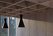 Retro pendant lamps with black metal lampshades suspended from modern coffered ceiling in pale larch wood and sunlight falling on wood-panelled wall