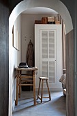 Arched doorway with view of rustic table, wooden stool and white cupboard with louver door