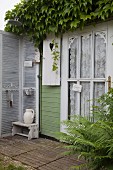 Ausschnitt eines Holzhäuschens mit berankter Hausfassade, an Tür Spitzenvorhang, kleine Terrasse mit Waschecke vor Paravent