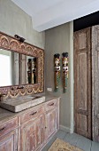 Vintage-Waschtisch mit Unterschrank an Wand, Ethno-Spiegel mit verziertem Holzrahmen und bemalte, asiatische Skulpturen an Wand