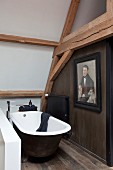 Free-standing vintage bathtub with designer floor-mounted taps below old roof structure with old portrait of man on dark brown wall