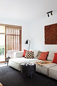 Moderne Wohnzimmerecke mit hellem Polstersofa und gemusterten Kissen, im Hintergrund Terrassentür mit Holzlamellen-Element