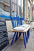 A bench made from three blue wooden chairs and a wooden plank in front of a restaurant