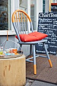 A newly painted grey and orange Windsor chair with an orange cushion in front of a restaurant