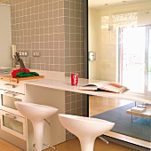 Kitchen counter with protruding breakfast bar and white bar stools in front of glass panels looking into living room