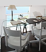 Glass tabletop on trestles and wooden swivel chairs in workspace on gallery; view of sea through glass balustrade