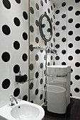 View through glass screen of white washstand with floor-mounted taps against black and white polka dot walls