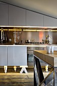 View past table to kitchen counter with glossy grey fronts, illuminated splashback and base units with unusual feet under sloping ceiling