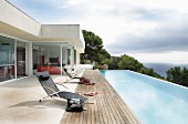 Contemporary holiday home with projecting flat roof; sun loungers on wooden deck next to infinity pool