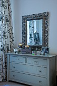 Simple, pale grey chest of drawers below mirror with carved frame
