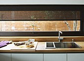 Eckige Edelstahlspüle in Küchenarbeitsfläche mit Naturholzplatte, darüber ein Fensterband mit transparentem Springrollo