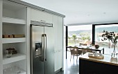 Stainless steel, side-by-side fridge freezer flanked by kitchen cabinets and shelving; dining area in front of panoramic window in background