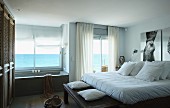 Double bed with white bed linen and bathtub below window with panoramic sea view