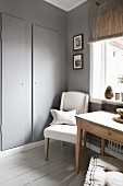 Küchentisch und gepolsterter Stuhl vor Fenster, daneben grau lackierte Wandschranktür