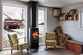 Fifties armchair next to fire in black, cast iron wood-burning stove in front of French windows with view of jetty