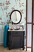 Dressing table with dark wooden base cabinet, pale top and oval mirror against leaf-patterned wallpaper