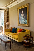 Yellow couch with colourful velvet scatter cushions and patterned footstool below antique, gilt-framed portait