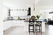 Open-plan white kitchen with black bar stools at counter and woman in lounge area in background to one side