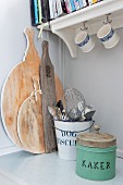 Vintage tin, metal bucket of kitchen utensils and wooden chopping boards on kitchen base unit below bracket shelf